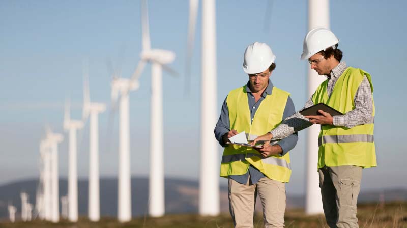 Engineers in safety vests and hard hats reviewing plans in front of a wind farm, highlighting Halker’s renewable energy engineering expertise in wind power development, site assessment, and sustainable infrastructure solutions.