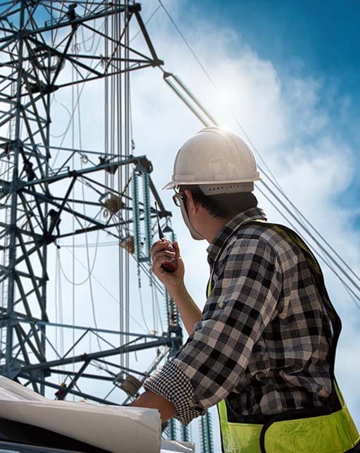 Field Engineer in hard hat and safety vest communicating by radio while reviewing plans near high-voltage transmission towers, highlighting Halker's expertise in power delivery engineering, grid infrastructure design, and utility construction oversight.