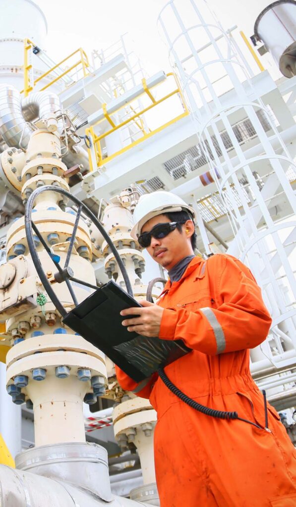 Field technician in safety gear inspecting industrial valves and piping with a handheld device, demonstrating Halker’s commitment to precise operations support, equipment reliability, and safe facility performance in energy and process environments.